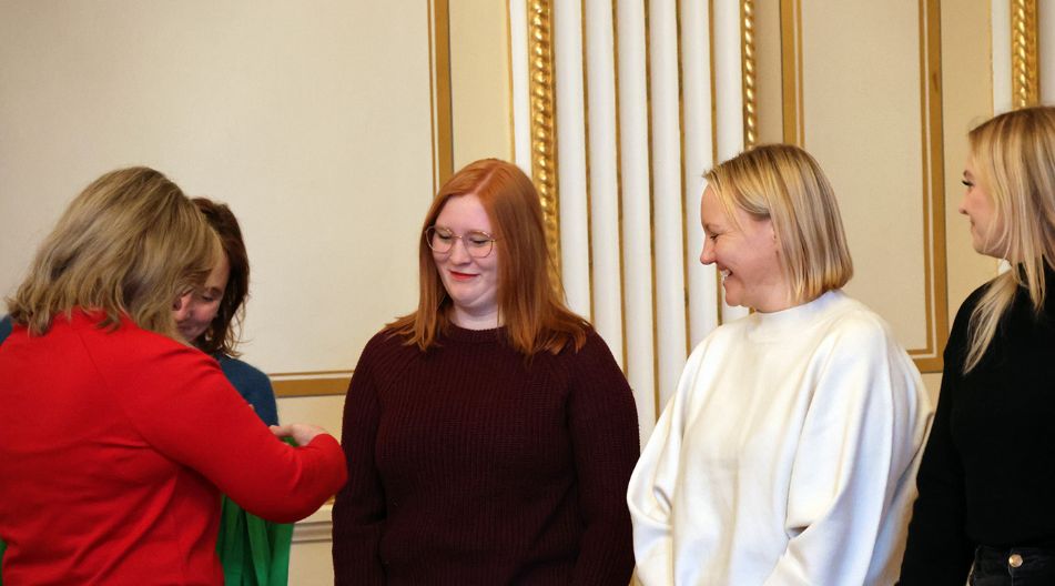 Mehrere Frauen stehen in einem Raum, eine überreicht oder befestigt einer anderen ein Abzeichen, die übrigen schauen lächelnd zu.