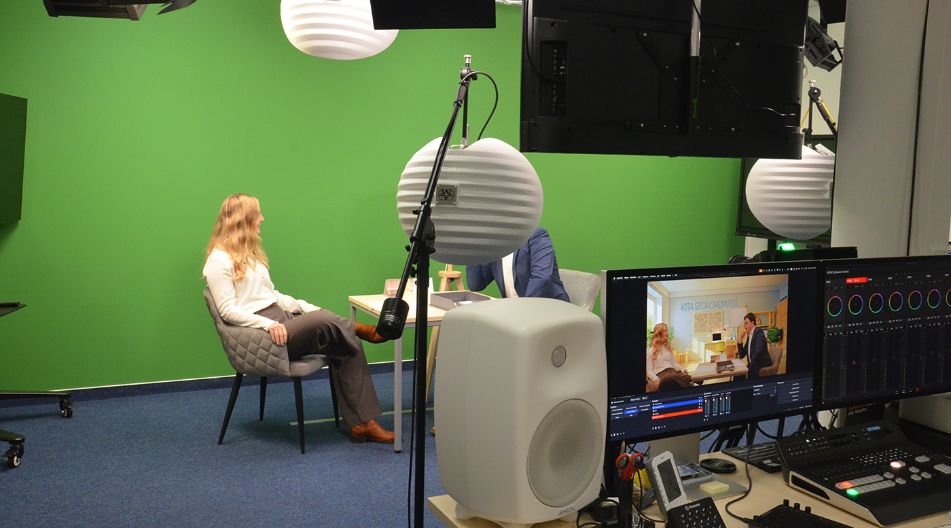 Zwei Personen sitzen sich in einem Studio mit grünem Hintergrund gegenüber, davor Monitor und technische Ausrüstung sichtbar