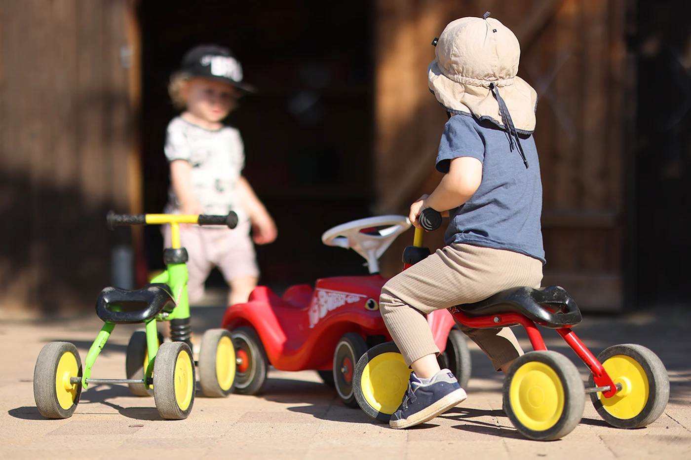 Ein Kind mit Sonnenhut sitzt auf einem roten Dreirad, während ein weiteres Kind mit Baseballkappe im Hintergrund steht. Mehrere bunte Kinderfahrzeuge stehen auf einem sonnigen Hof. Ein Kind mit Sonnenhut sitzt auf einem roten Dreirad, während ein weiteres Kind mit Baseballkappe im Hintergrund steht. Mehrere bunte Kinderfahrzeuge stehen auf einem sonnigen Hof.