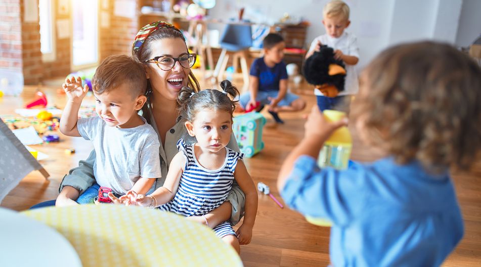 Ein heller und farbenfroher Kindergartenraum mit spielenden Kindern. Eine lachende Erzieherin sitzt mit zwei kleinen Kindern auf dem Boden, während andere Kinder um sie herum mit Spielzeug beschäftigt sind. Ein heller und farbenfroher Kindergartenraum mit spielenden Kindern. Eine lachende Erzieherin sitzt mit zwei kleinen Kindern auf dem Boden, während andere Kinder um sie herum mit Spielzeug beschäftigt sind.