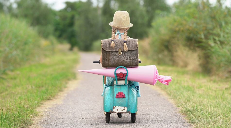 Ein Mädchen fährt auf einem Roller mit ihrer Schultüte einen Weg entlang. Ein Mädchen fährt auf einem Roller mit ihrer Schultüte einen Weg entlang.