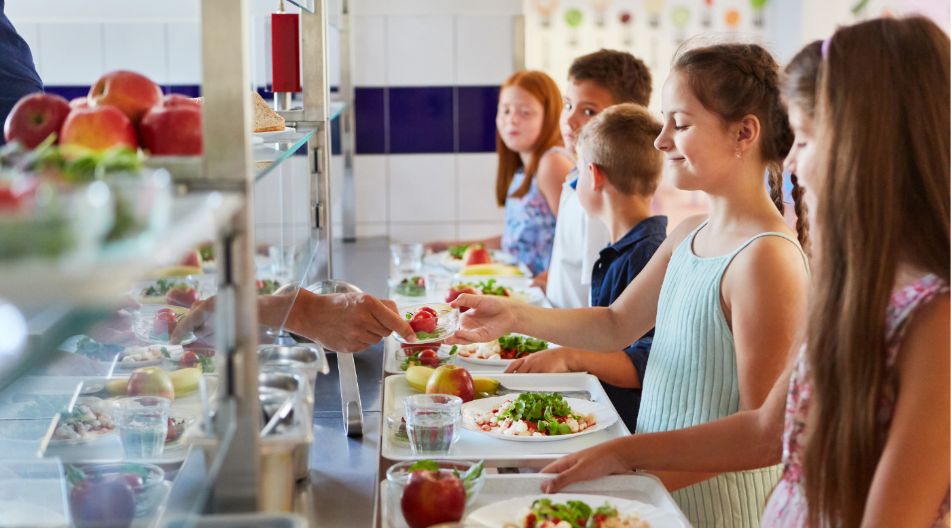 Kinder stehen in einer Schulkantine in der Schlange und bekommen gesundes Essen auf ihre Tabletts serviert.