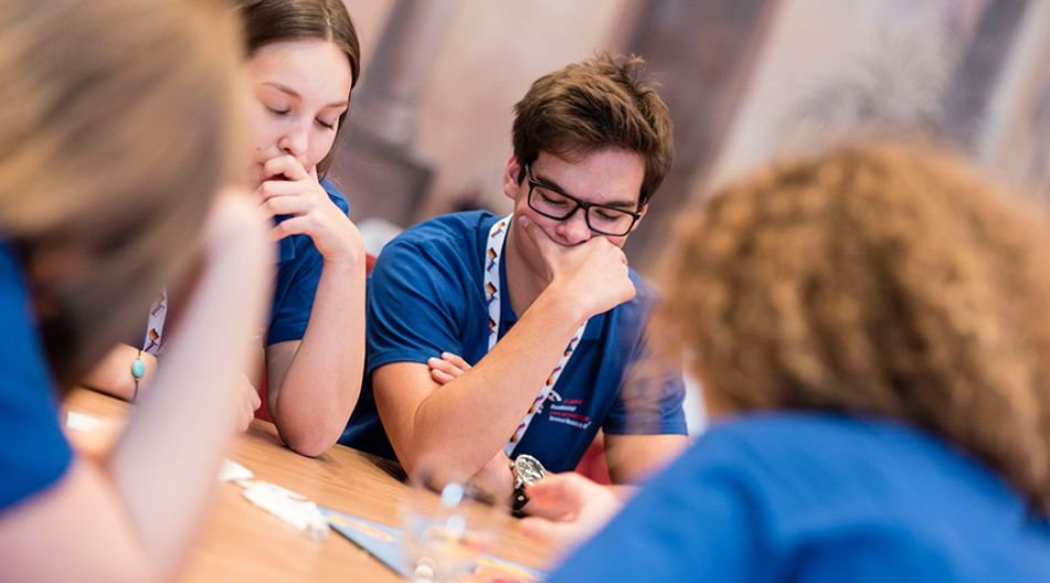 Wettkämpfe sind ein weiterer Baustein in der Begabungsförderung. Schülerinnen und Schüler sitzen an einem Spieltisch und denken nach.